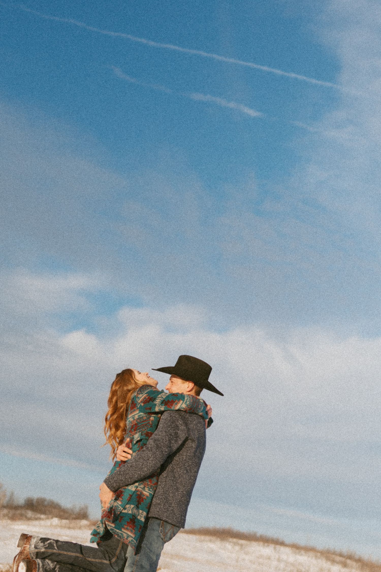 A romantic couple embraces in a snowy winter landscape beneath a bright blue sky.
