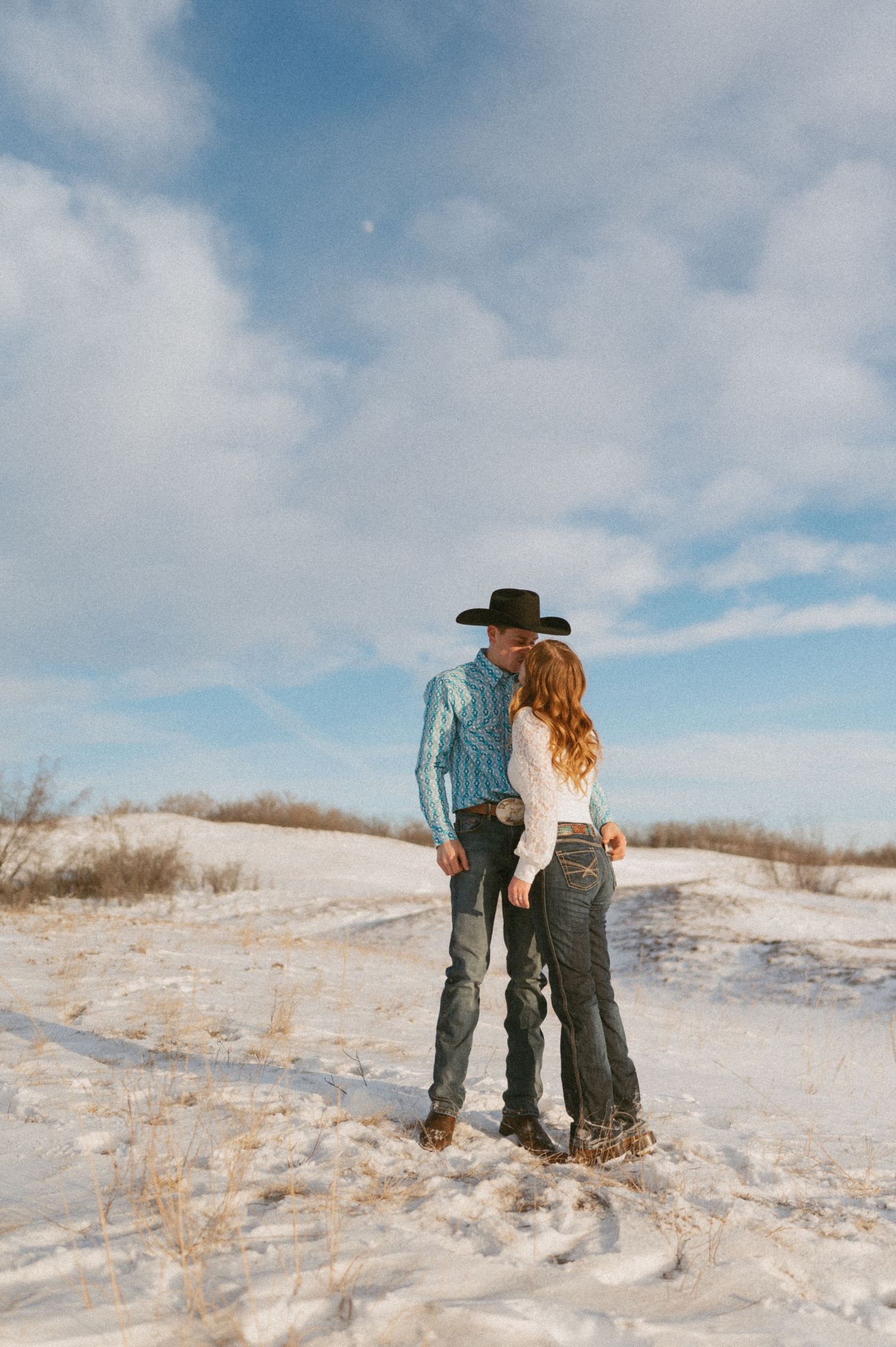 A couple stands together on a snow-covered hill against a cloudy blue sky.