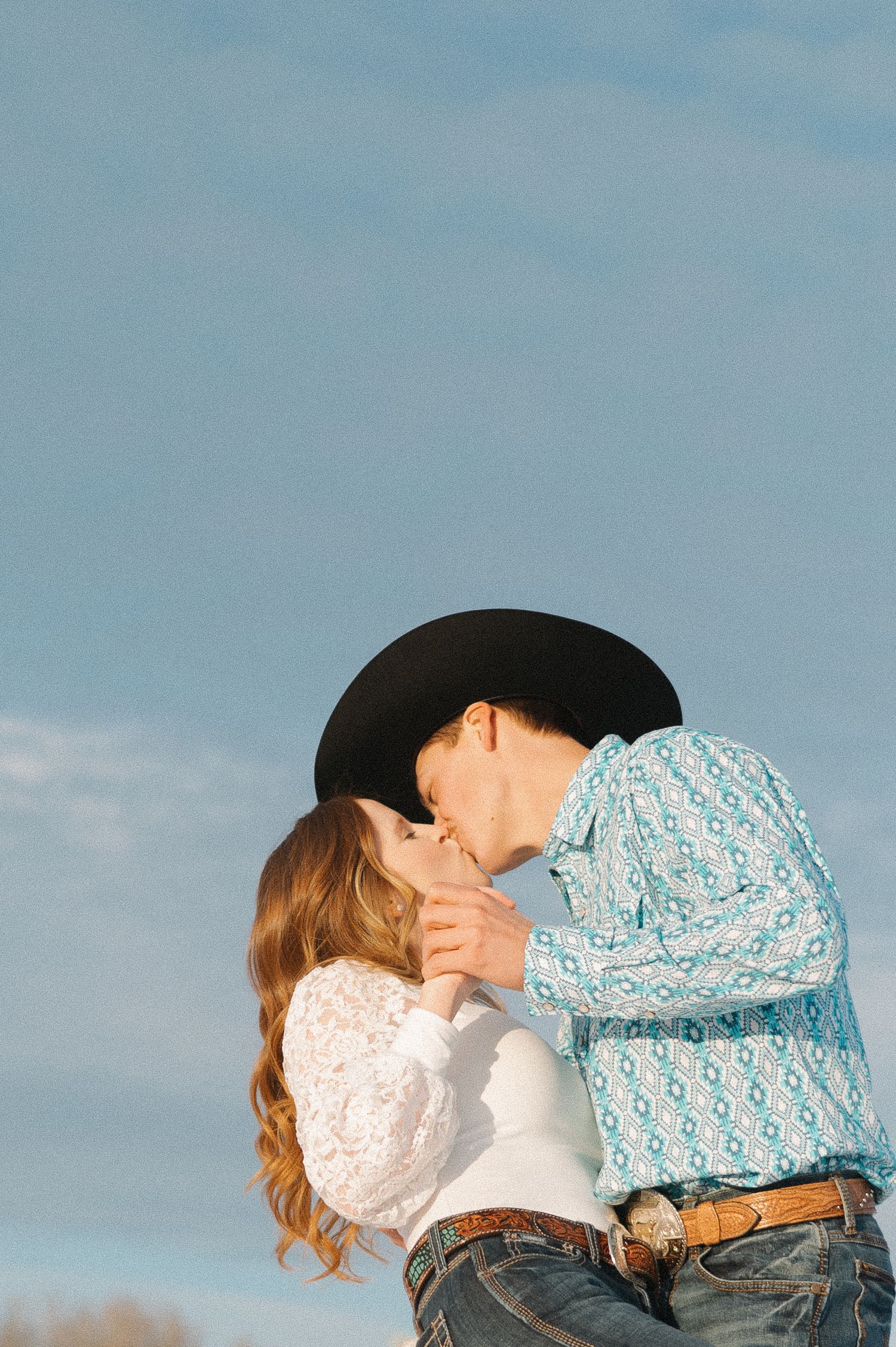 A romantic couple shares an intimate moment in western attire against a blue sky.