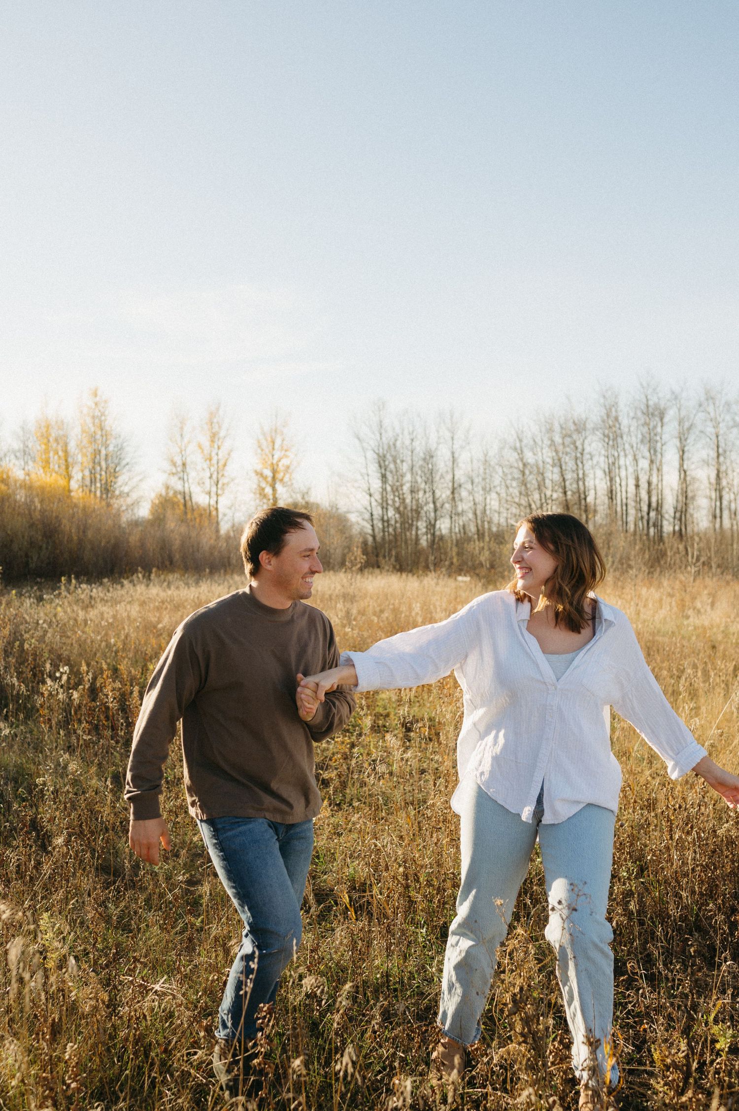Couple laughing and walking together through a golden sunlit field with trees in the background.