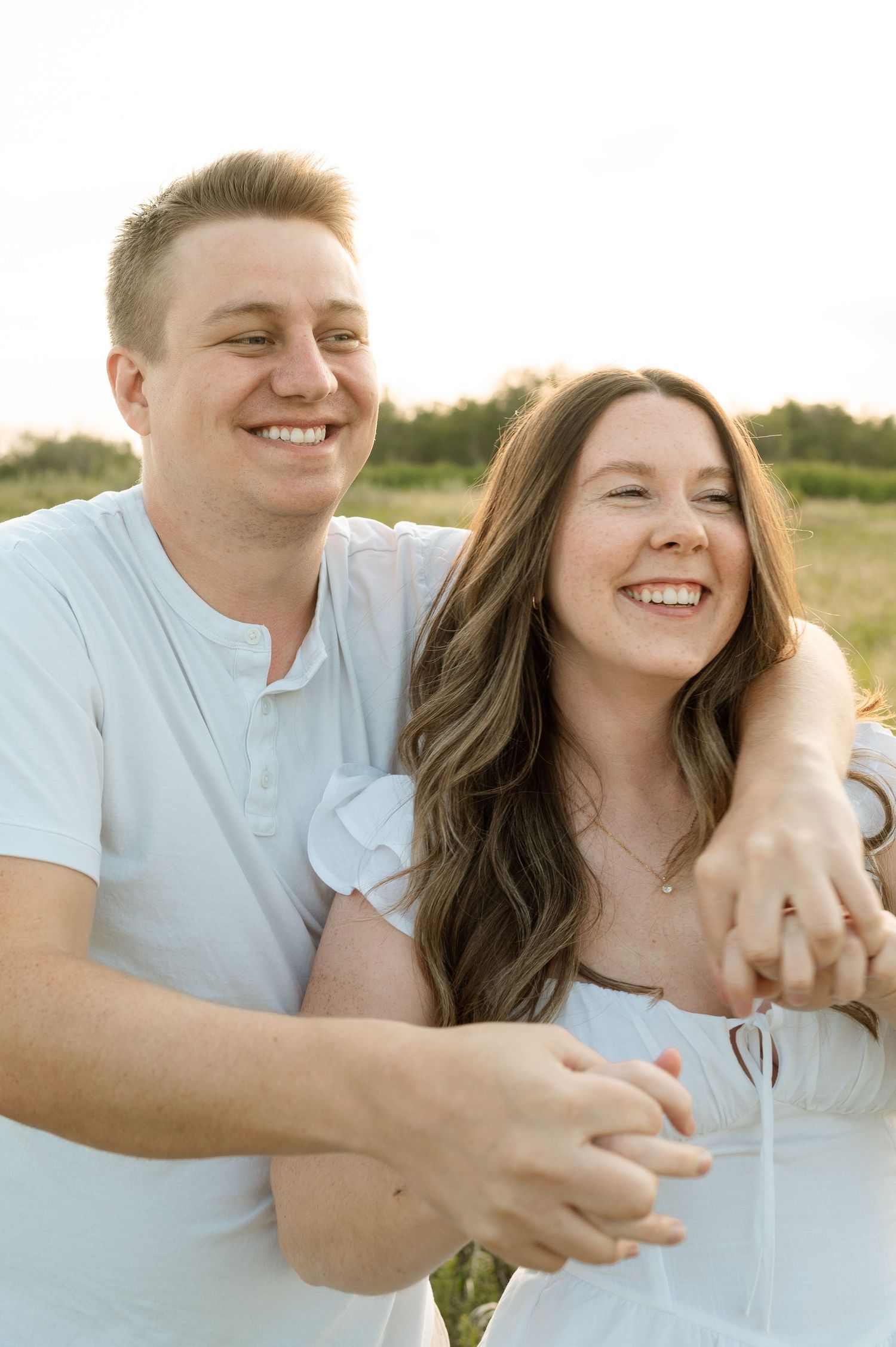 A couple shares a joyful laugh while holding hands during an outdoor sunset photoshoot in a field.