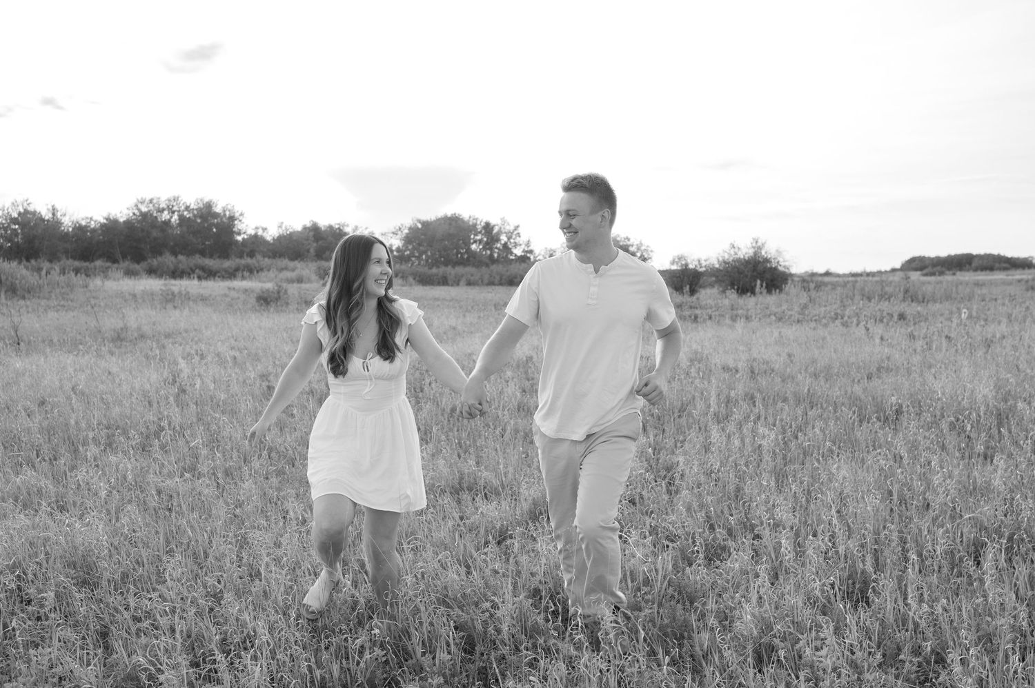 A couple stands together in a sun-dappled meadow or field wearing white outfits during what appears to be an engagement shoot.