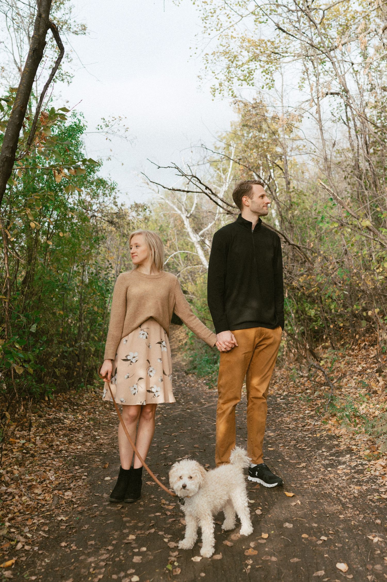 Couple walking small white dog on autumn trail while holding hands.