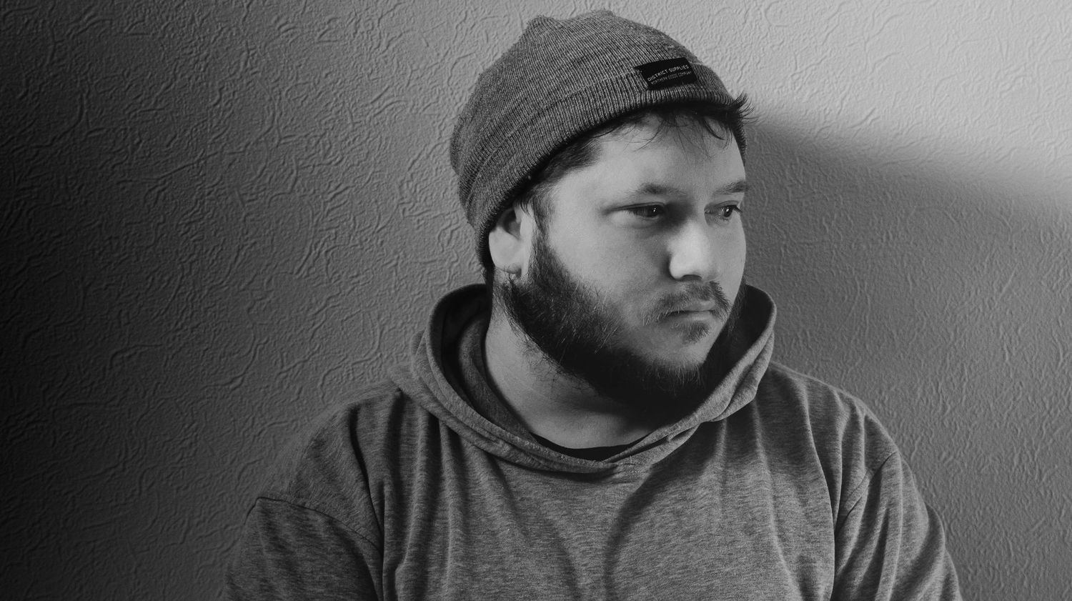 Black and white portrait of person wearing a beanie and hoodie looking to the side against a light wall.