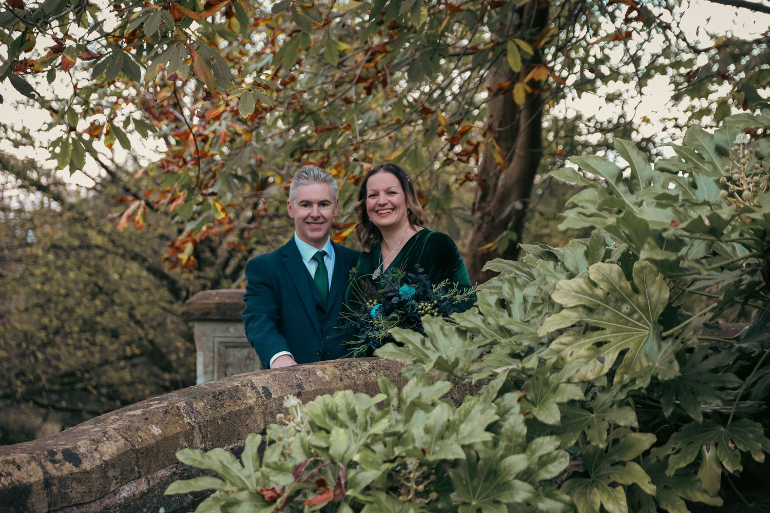 A couple stands together among autumn foliage in a garden setting.
