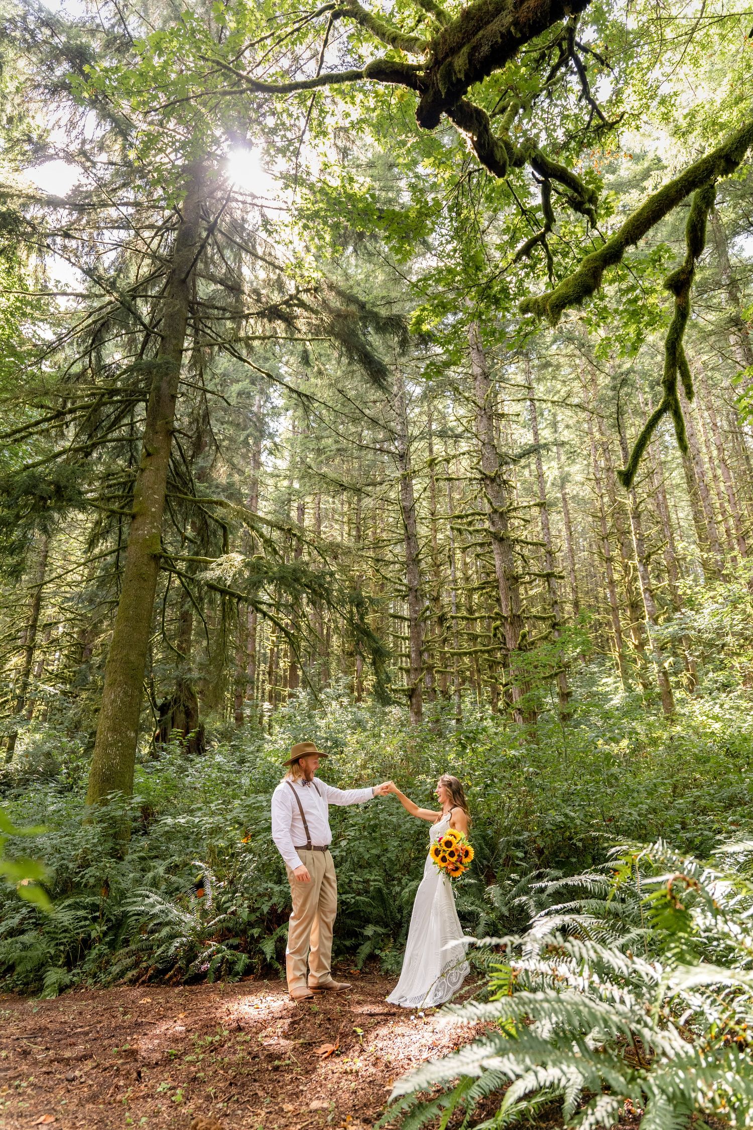 Couple in wedding attire holds hands in a sunlit Pacific Northwest forest with tall evergreen trees and ferns surrounding them.