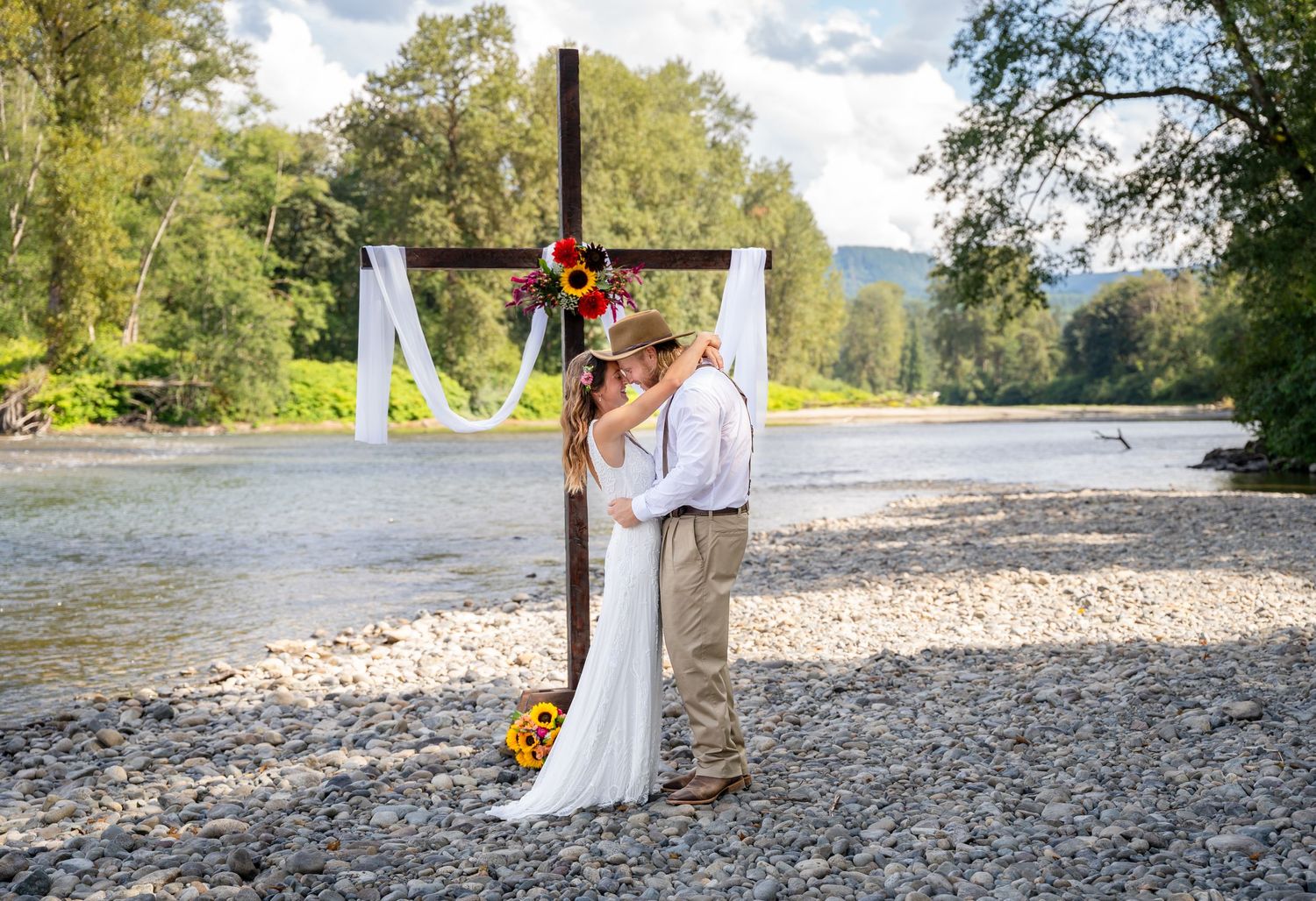 A couple embraces in a romantic outdoor wedding ceremony by a wooden cross adorned with sunflowers and white fabric.