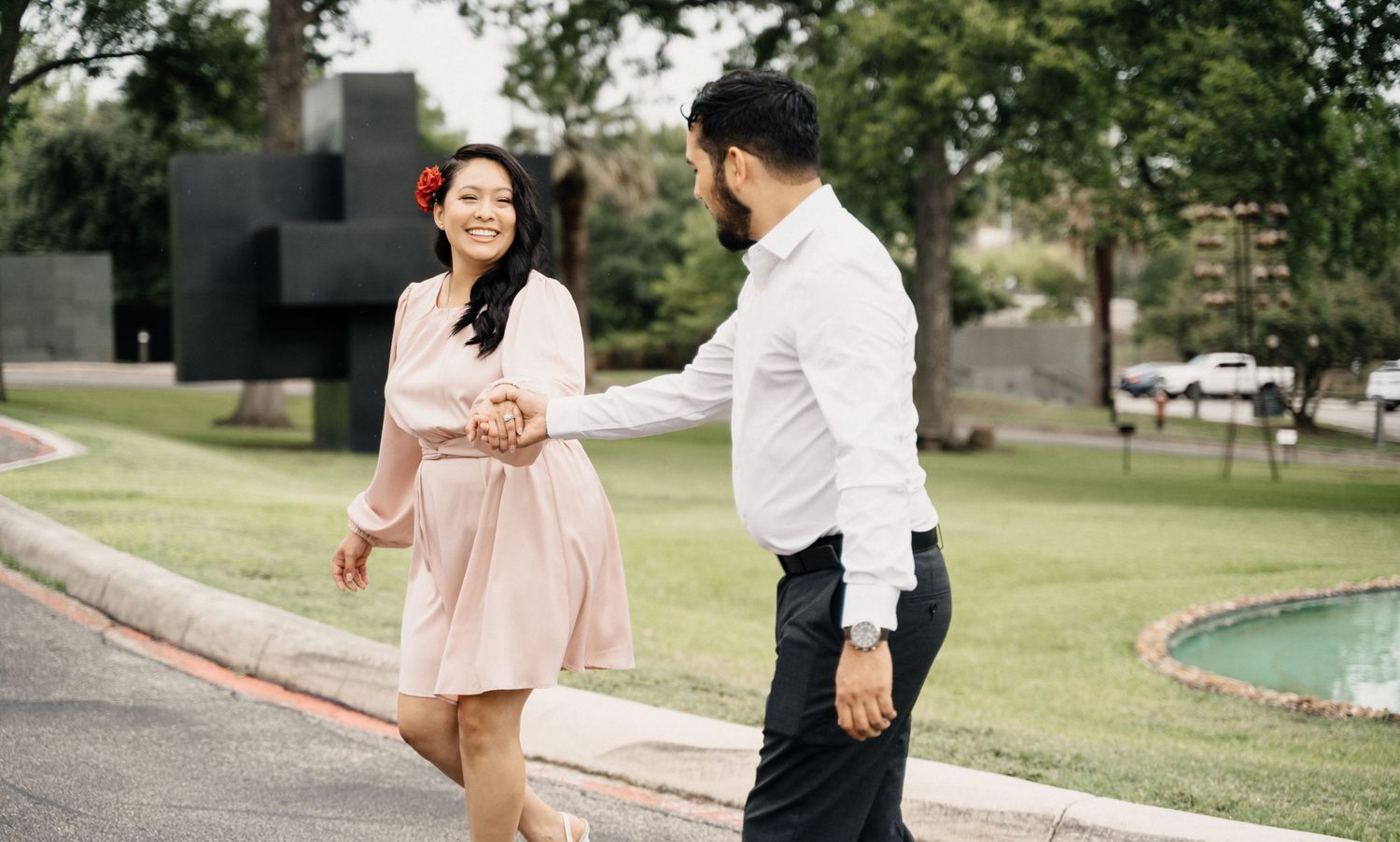 Couple in light-colored outfits holding hands and dancing together in a park with green trees in the background.