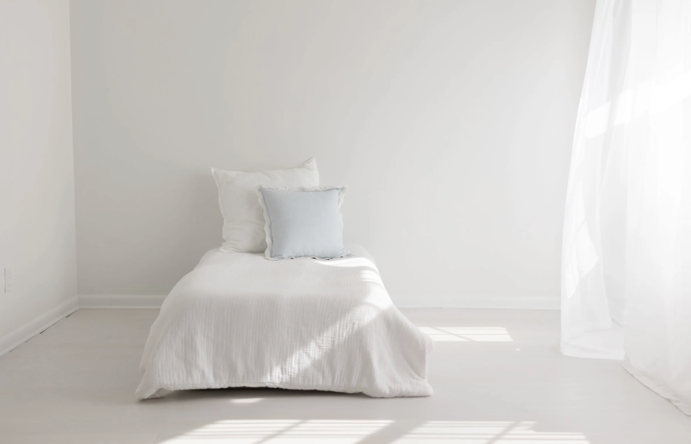 Minimalist white bedroom with bed, pillows and natural light streaming through sheer curtains.