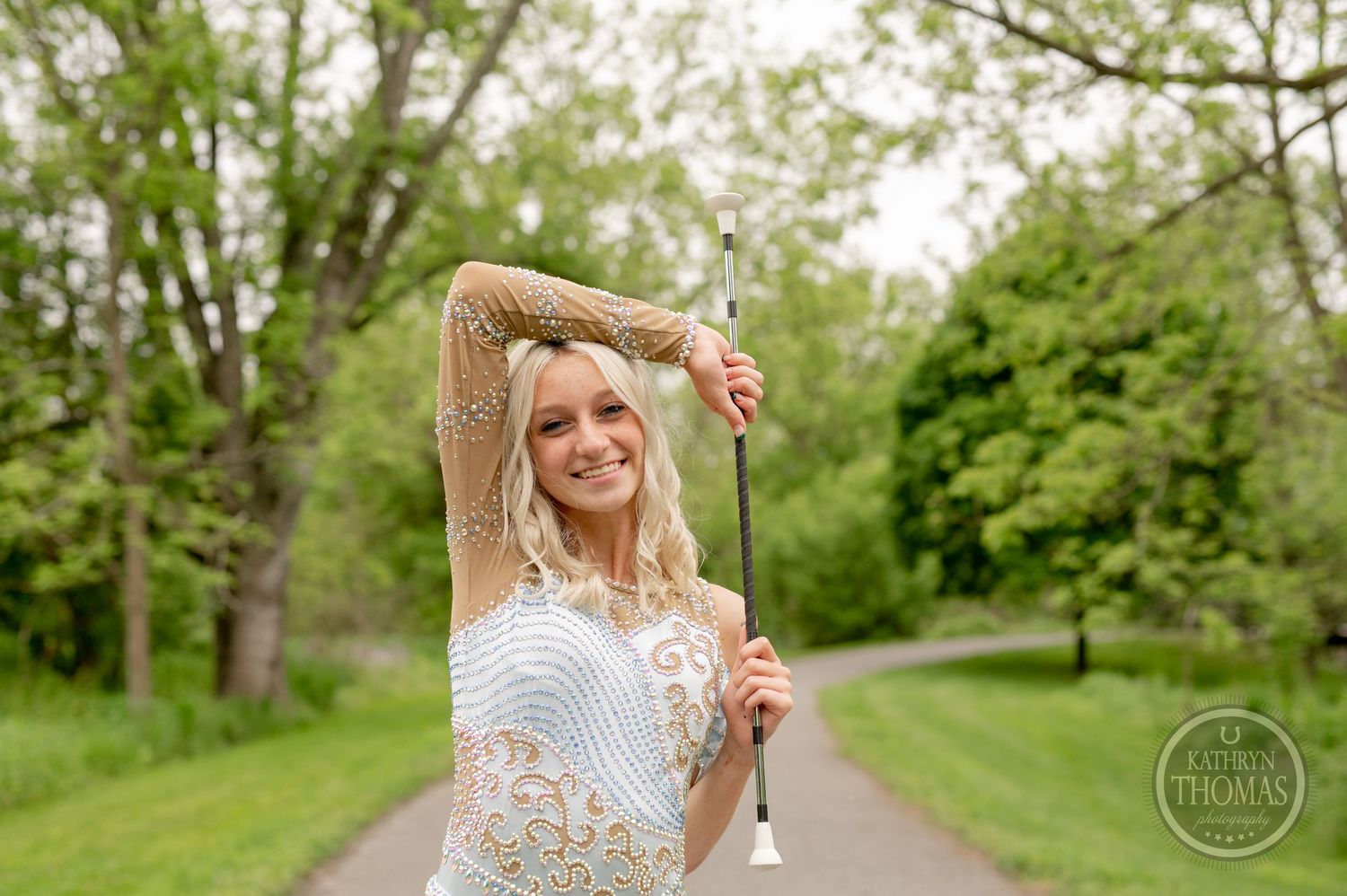 Contestant in a delicate lace dress strikes a dynamic pose on a tree-lined path.
