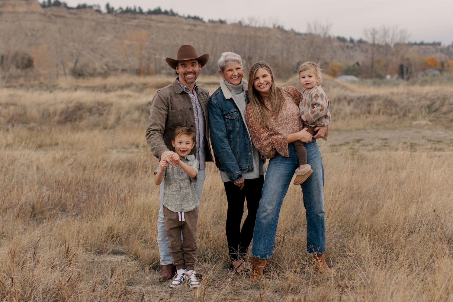 A rustic family portrait in autumn featuring five people standing together in a golden field with rolling hills behind them.