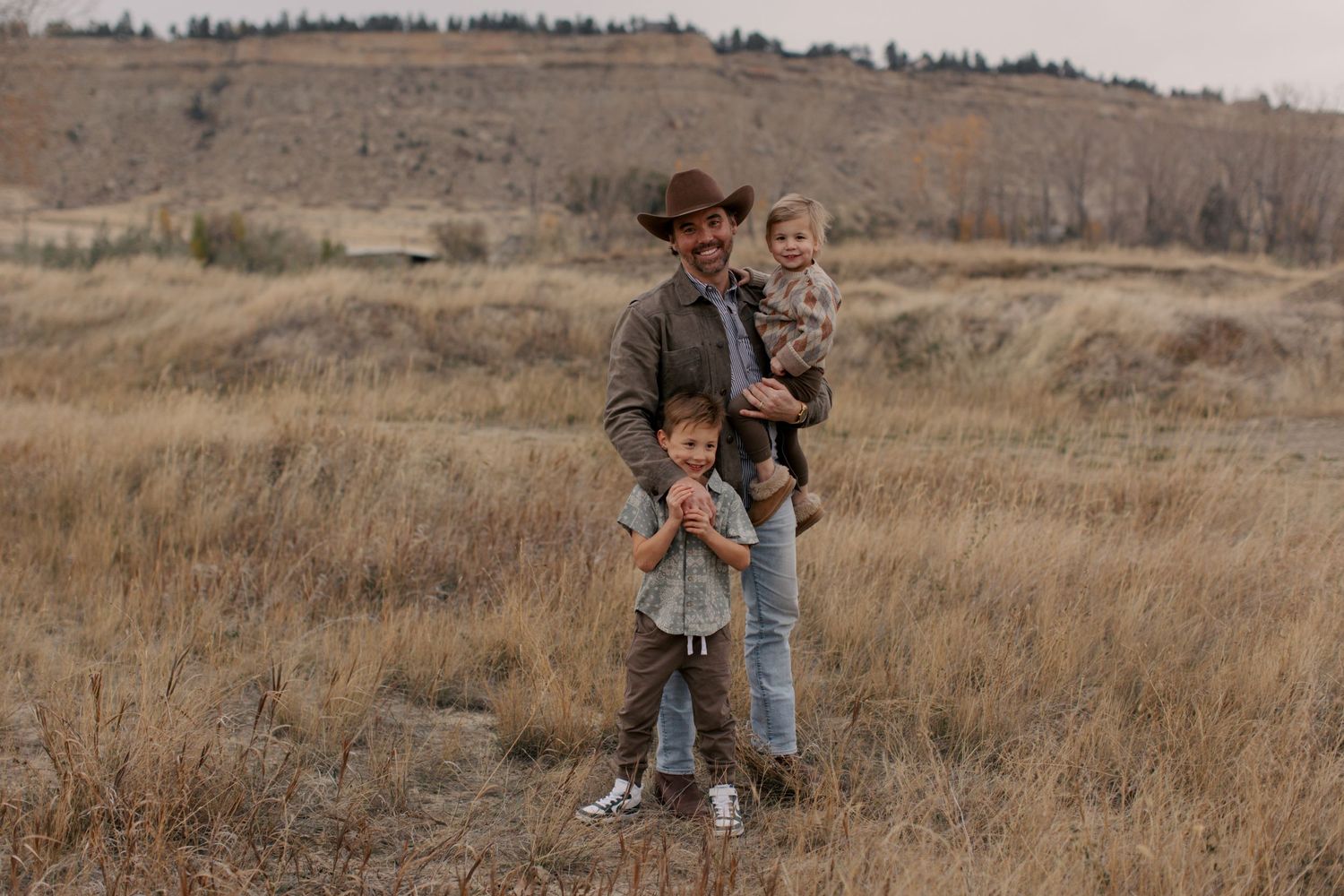 Three people stand together in brown autumn grassland with rolling hills in the background.