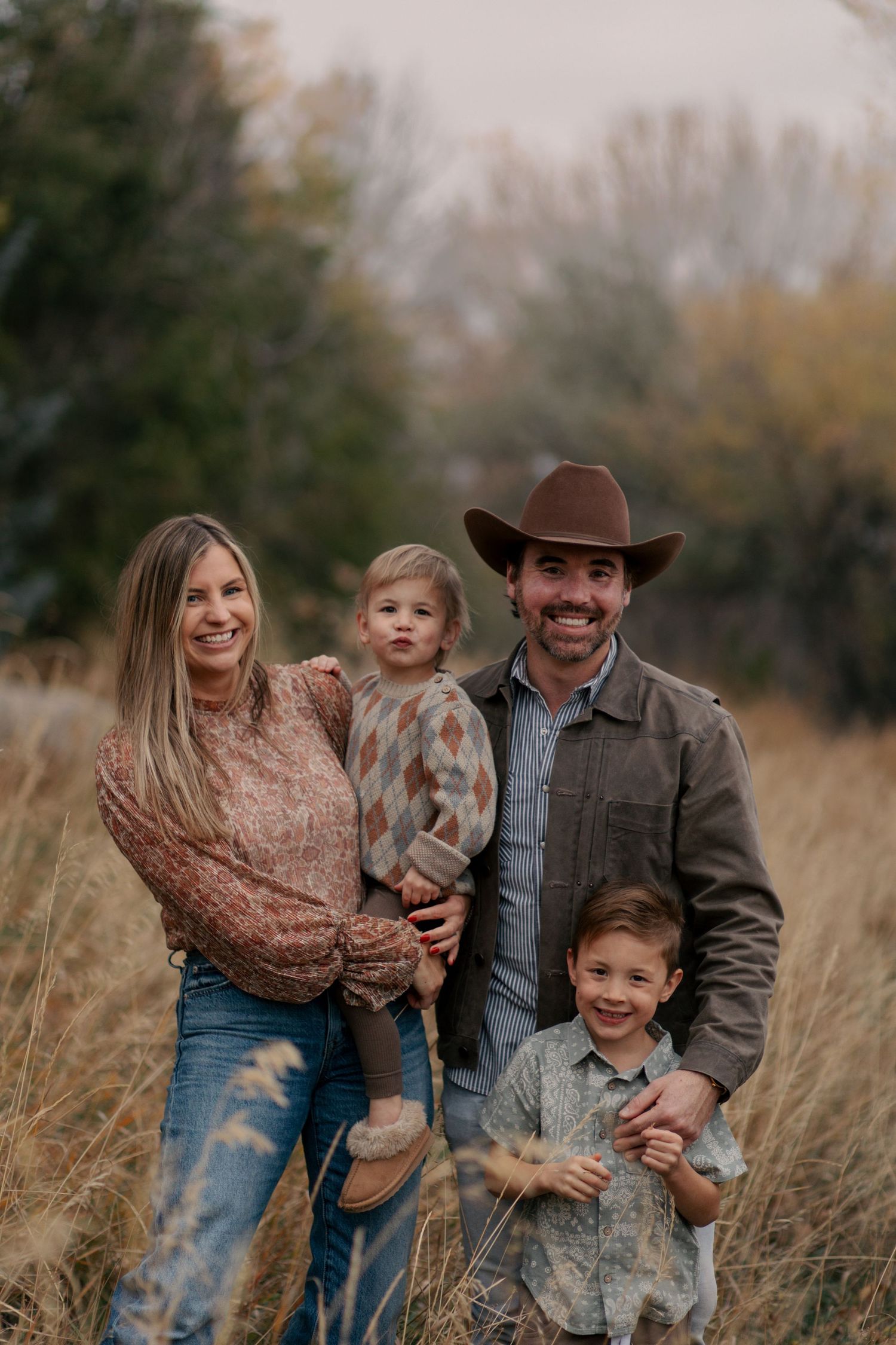 A family poses together in a field of tall grass during autumn with a brown cowboy hat and casual attire.