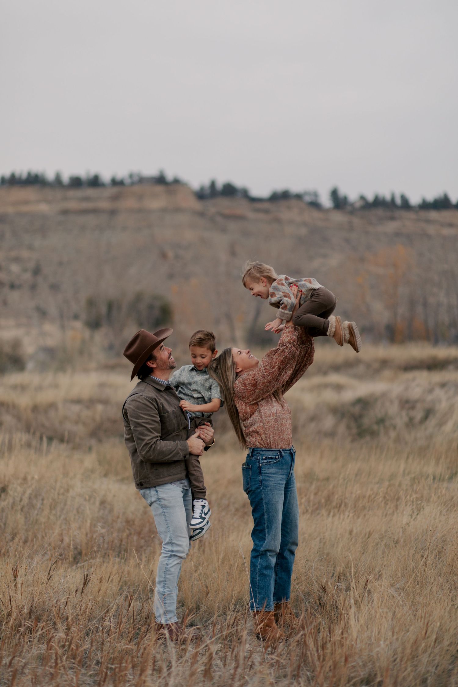 A couple lifts their children playfully in a grassy field with rocky cliffs in the background on an overcast day.