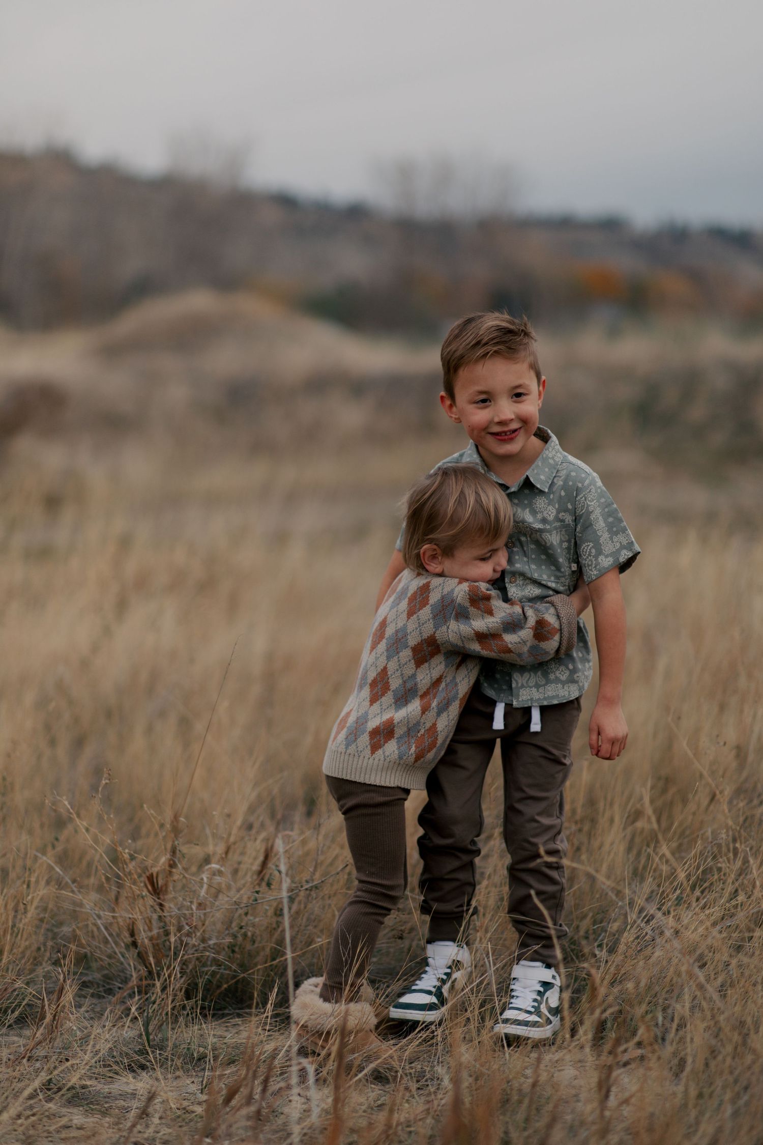Two children stand together hugging in a field of tall dry grass during an overcast day.