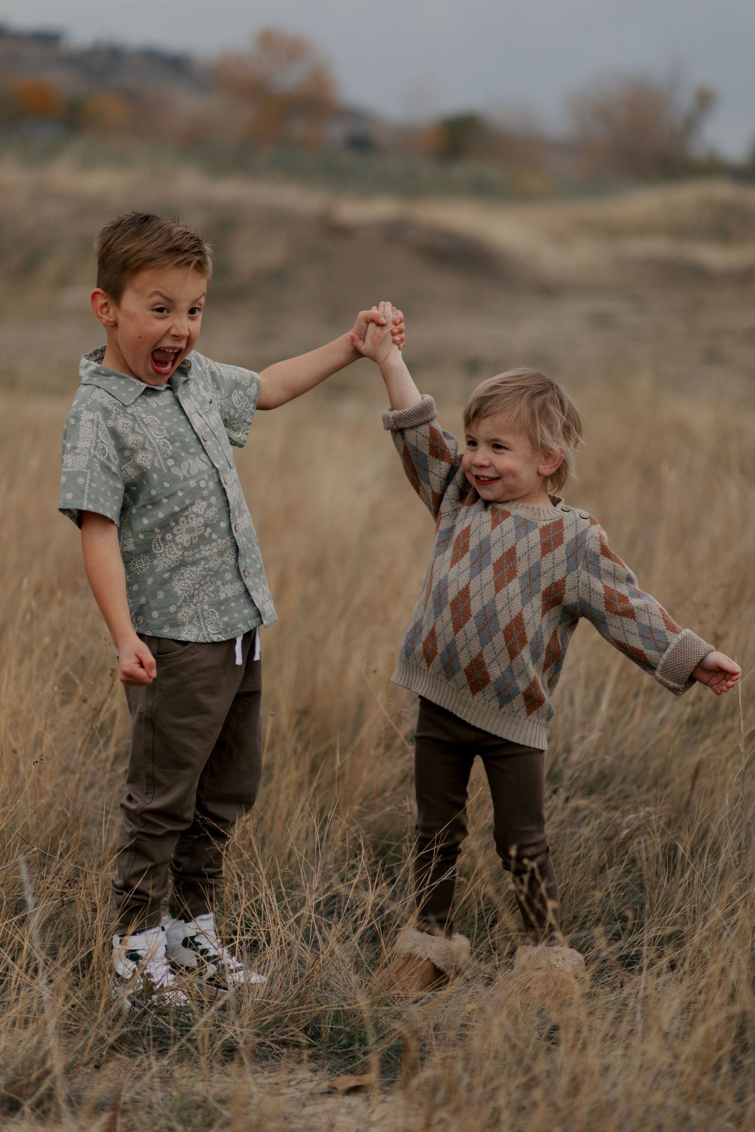 Two children hold hands and laugh while playing together in a field of tall brown grass on an autumn day.