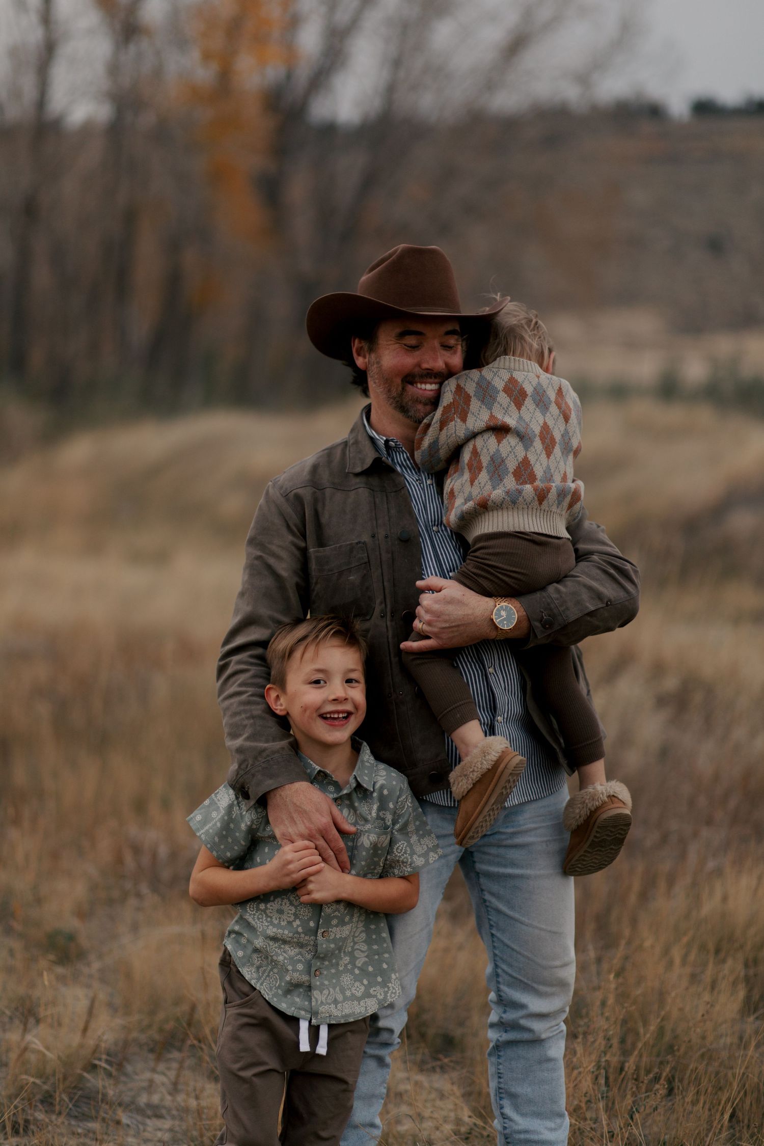 A person in a cowboy hat and jacket shares a joyful moment with children in an autumn field at sunset.