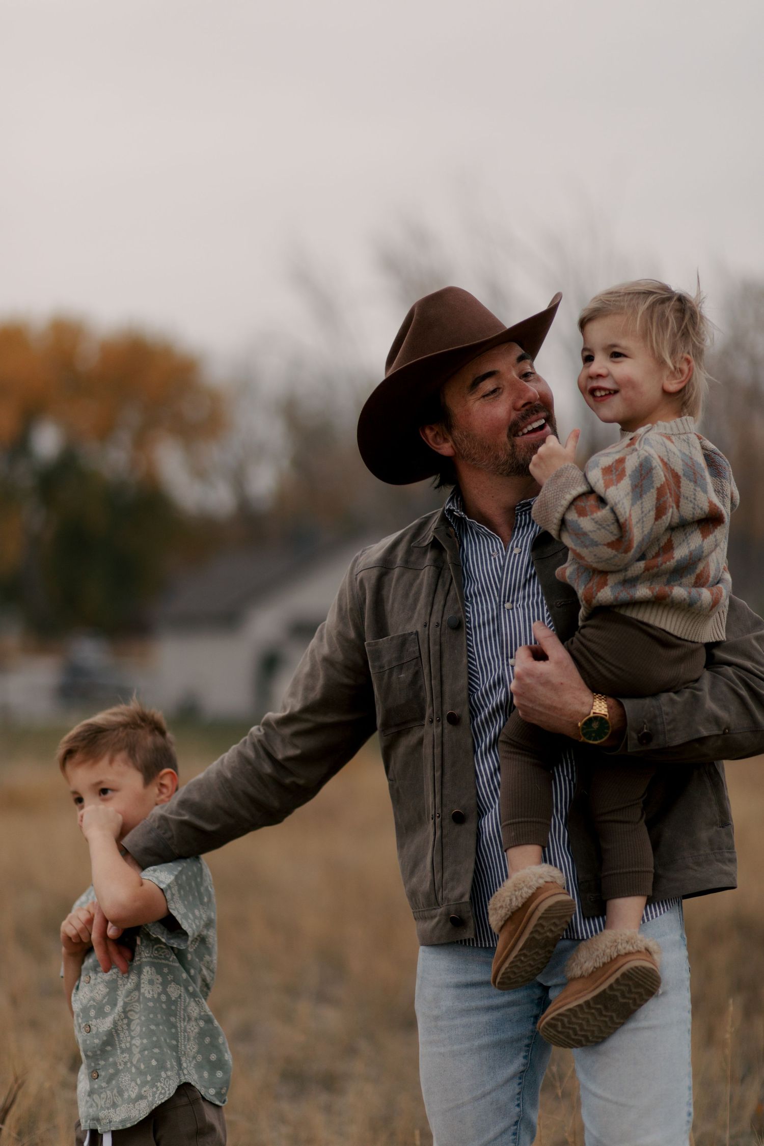 A grown man in a cowboy hat and denim jacket shares a joyful moment with two children outdoors at dusk.