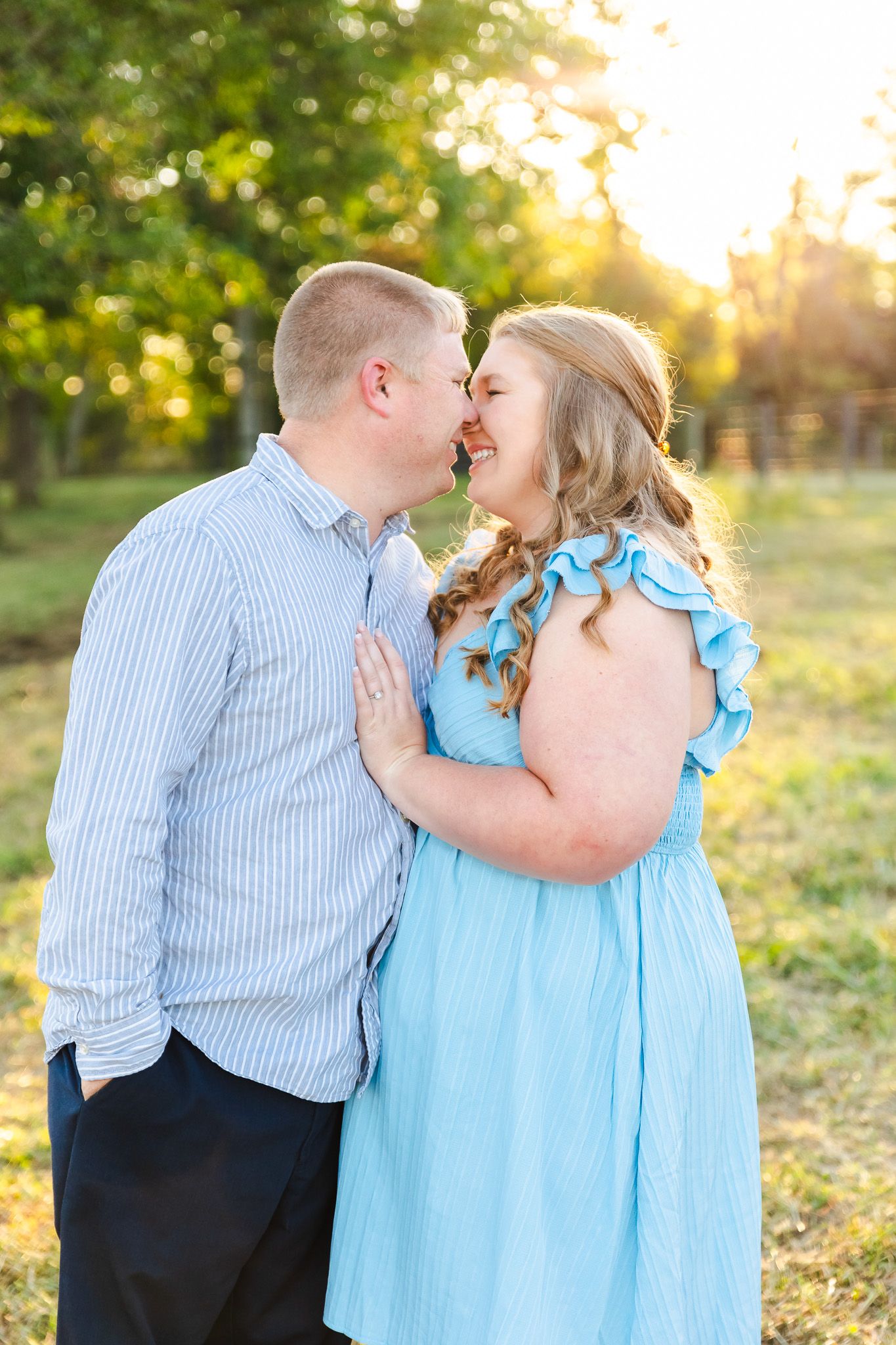 Romantic outdoor photoshoot at sunset with couple in blue and white outfits embracing in a grassy field.