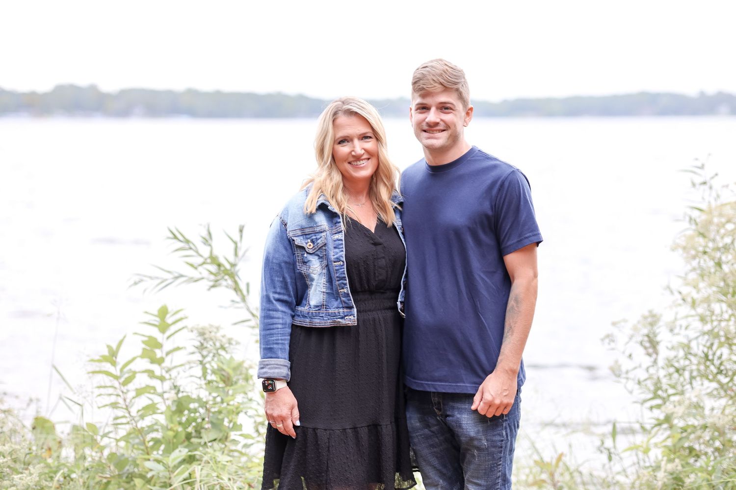 A mother and son together smiling by a waterfront with trees and vegetation in the background on an overcast day.
