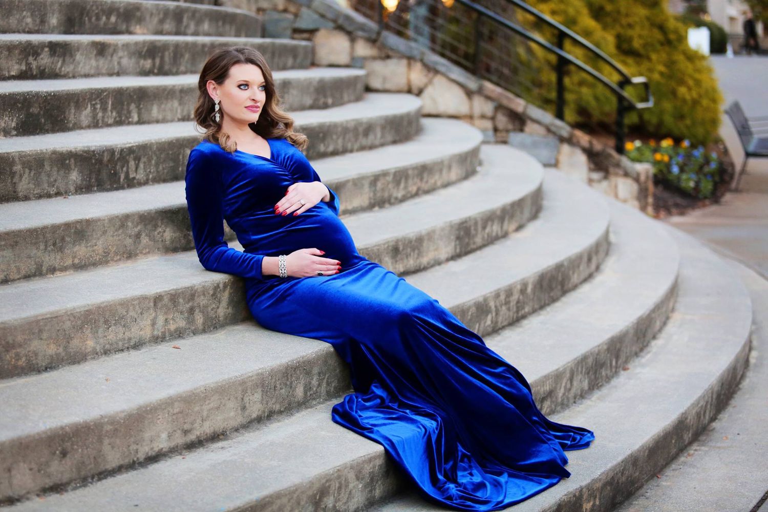 A maternity portrait of a woman in royal blue velvet dress reclining on concrete steps.