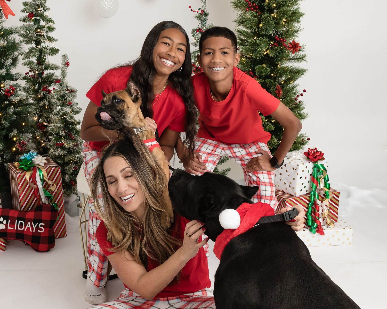 A family in red holiday attire poses with their black dogs on a white backdrop with Christmas trees and presents.