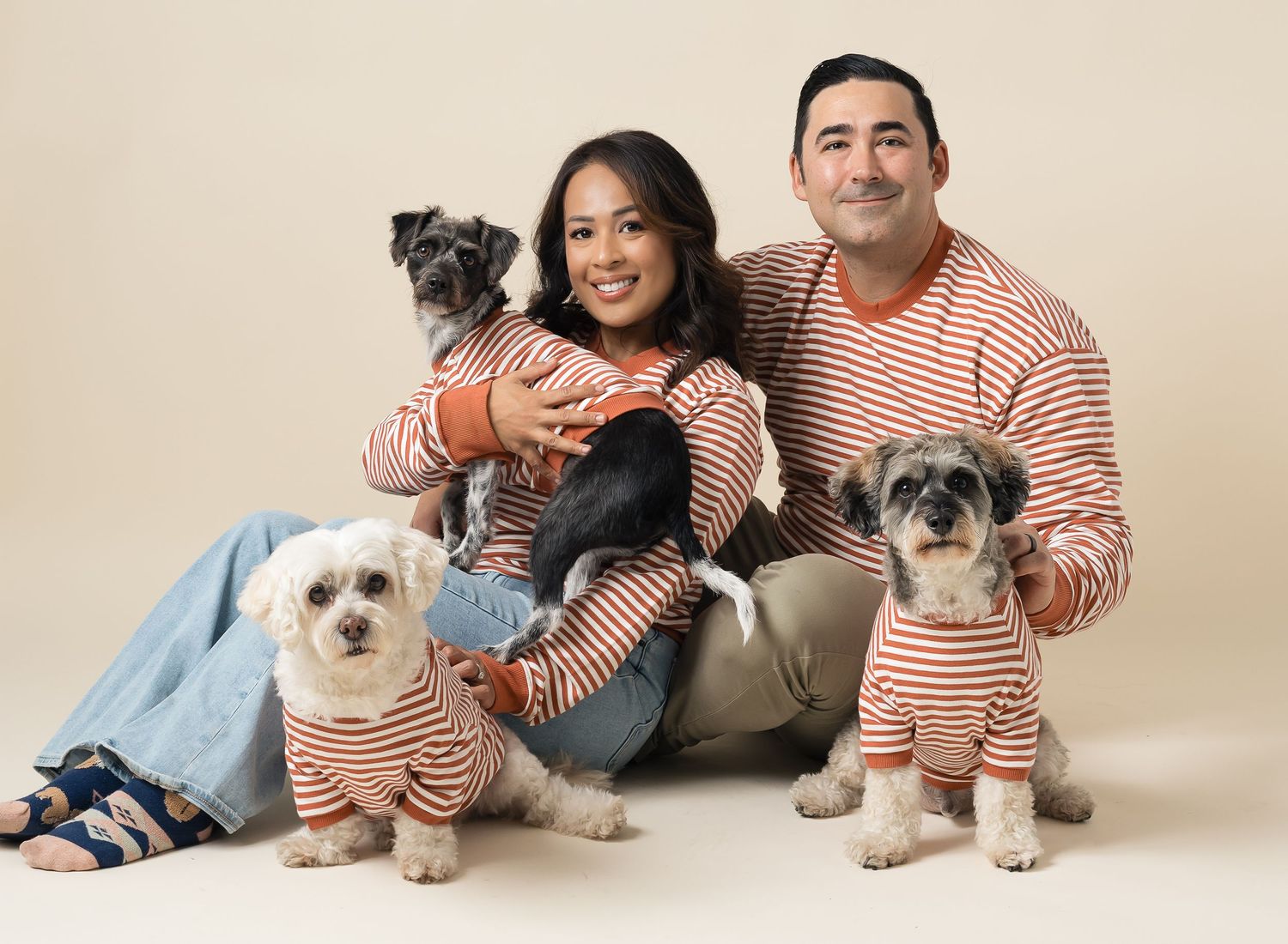 A family wearing matching striped shirts poses with their small white dog on a light colored backdrop.