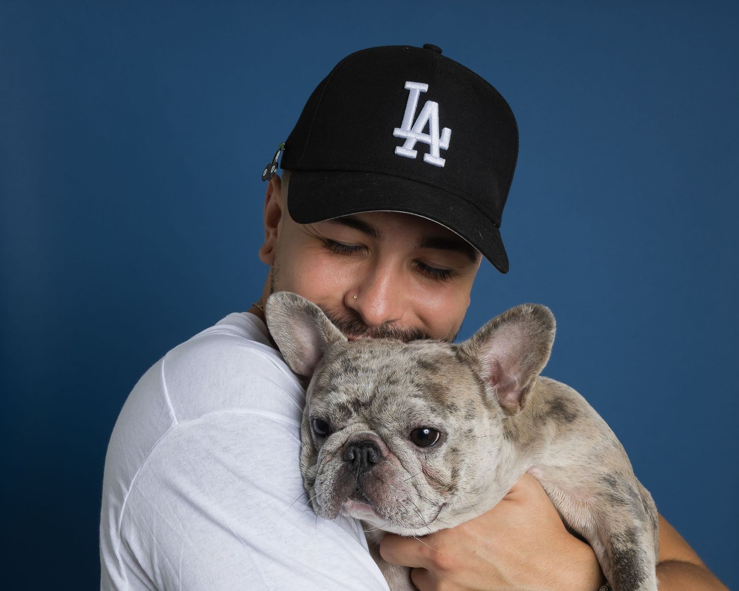French bulldog being held and cuddled against dark blue backdrop while wearing LA baseball cap.