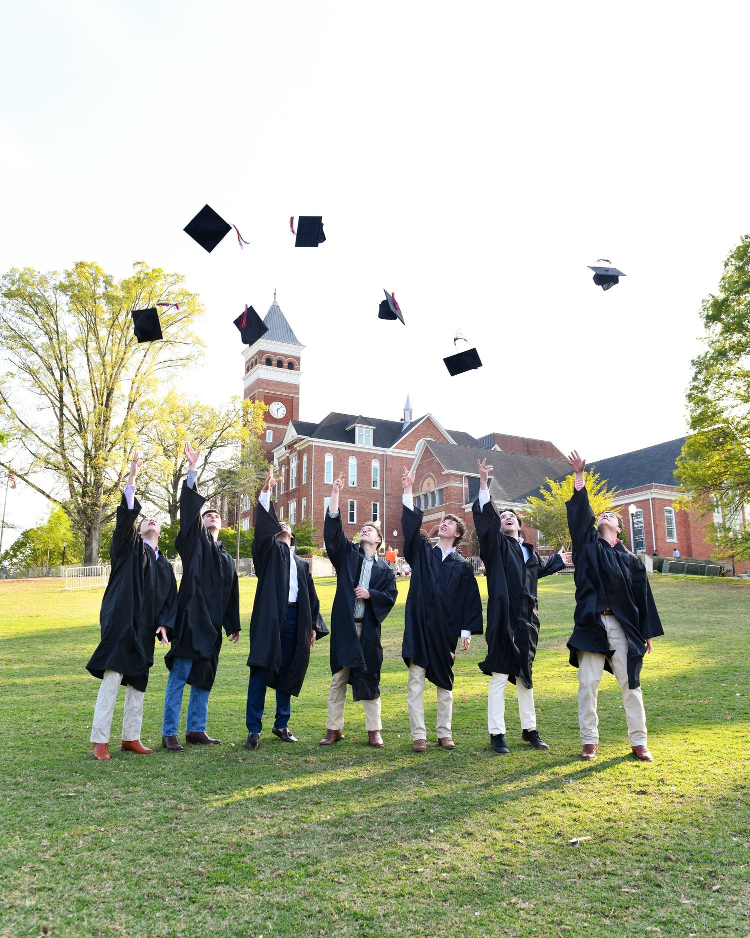 Graduates throw their caps in the air on lawn in front of brick university building with tower.
