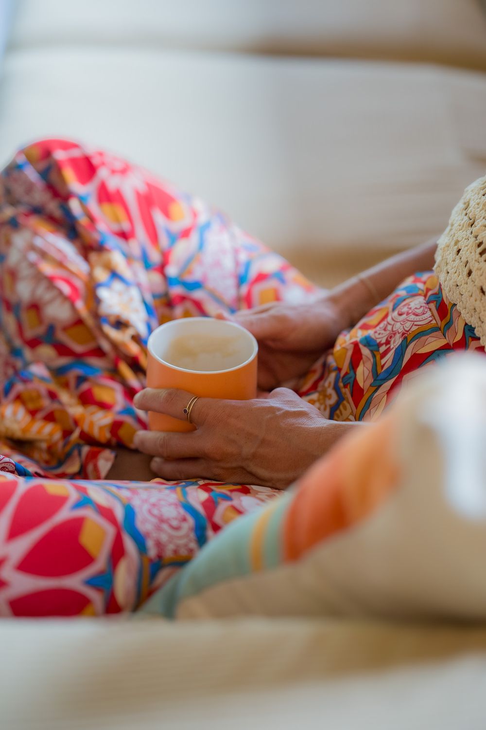 Hands holding a white coffee cup while wearing colorful floral patterned pajamas or loungewear on a cozy couch.