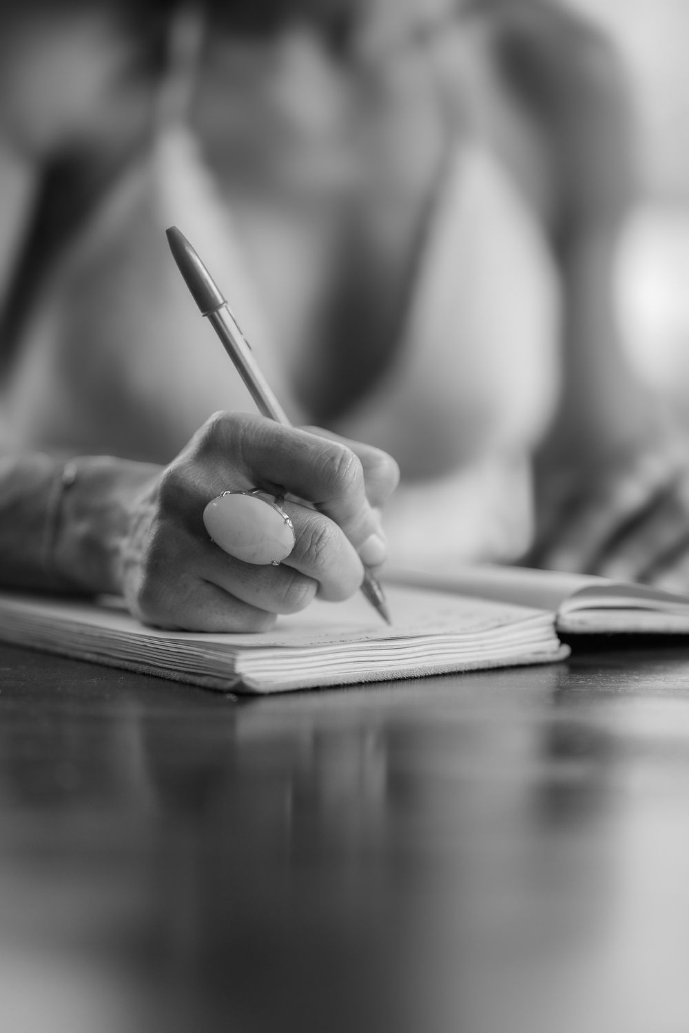 A close-up black and white sequence of hands writing with a pen in a notebook on a reflective surface.
