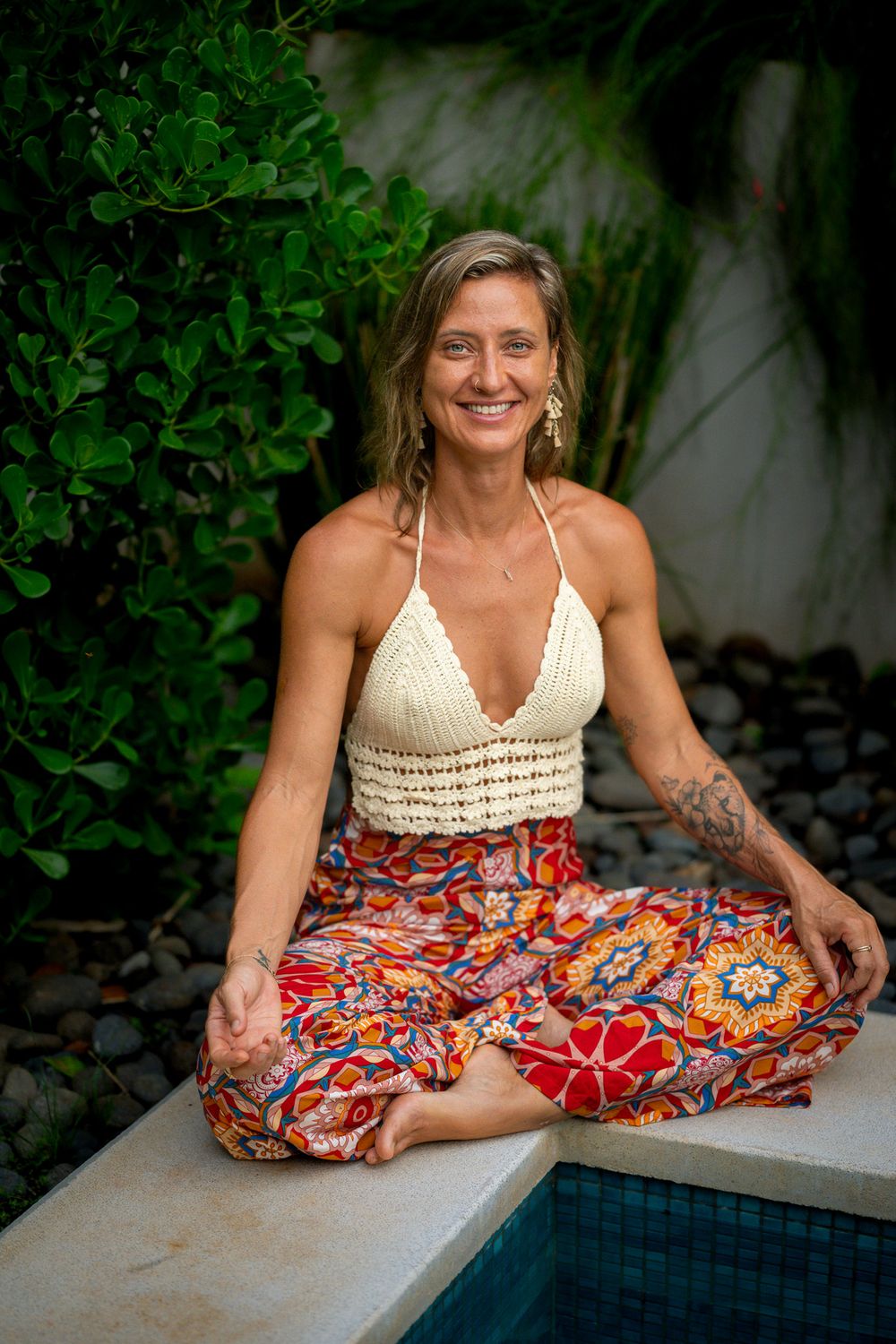 A person in a white crochet top and colorful floral skirt poses gracefully by a pool in a tropical garden setting.