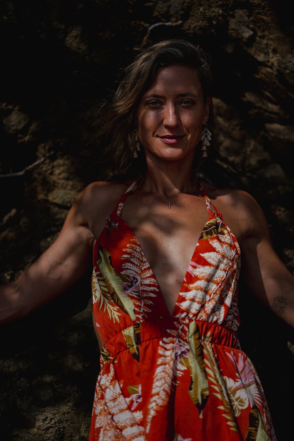 Person wearing vibrant red tropical print dress poses against rocky backdrop in dramatic lighting.
