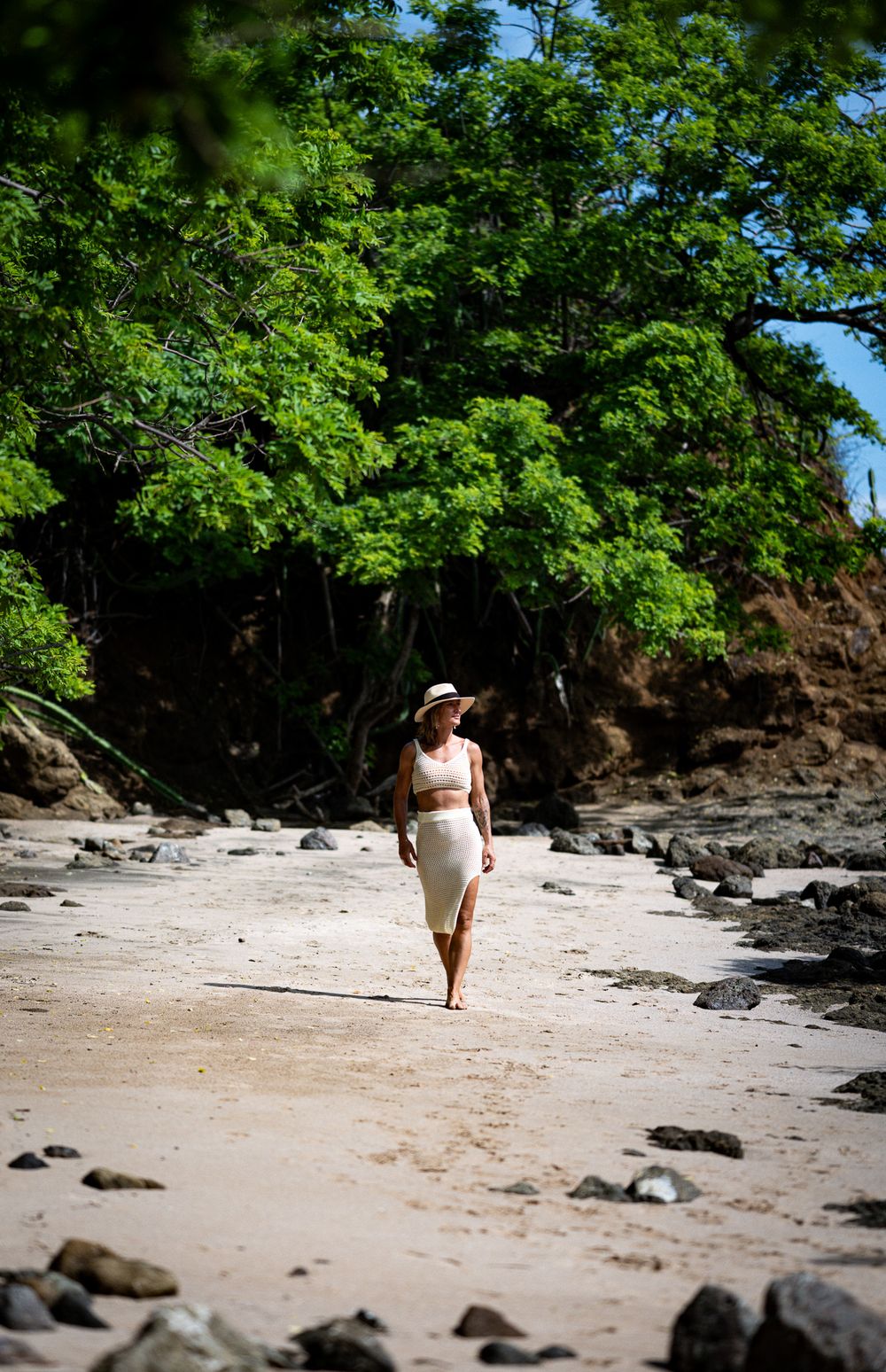 A person in white walks along a secluded beach with lush green trees and rocky outcroppings in the background.
