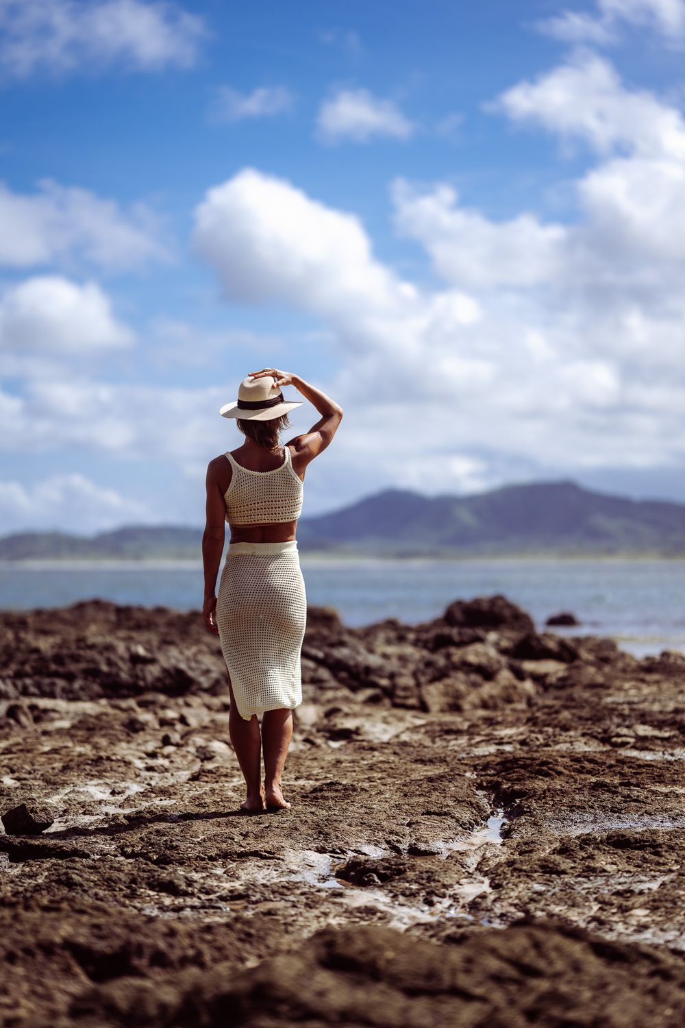 A person in white dress and sun hat walks along a rocky coastal shoreline on a sunny day with mountains in background.