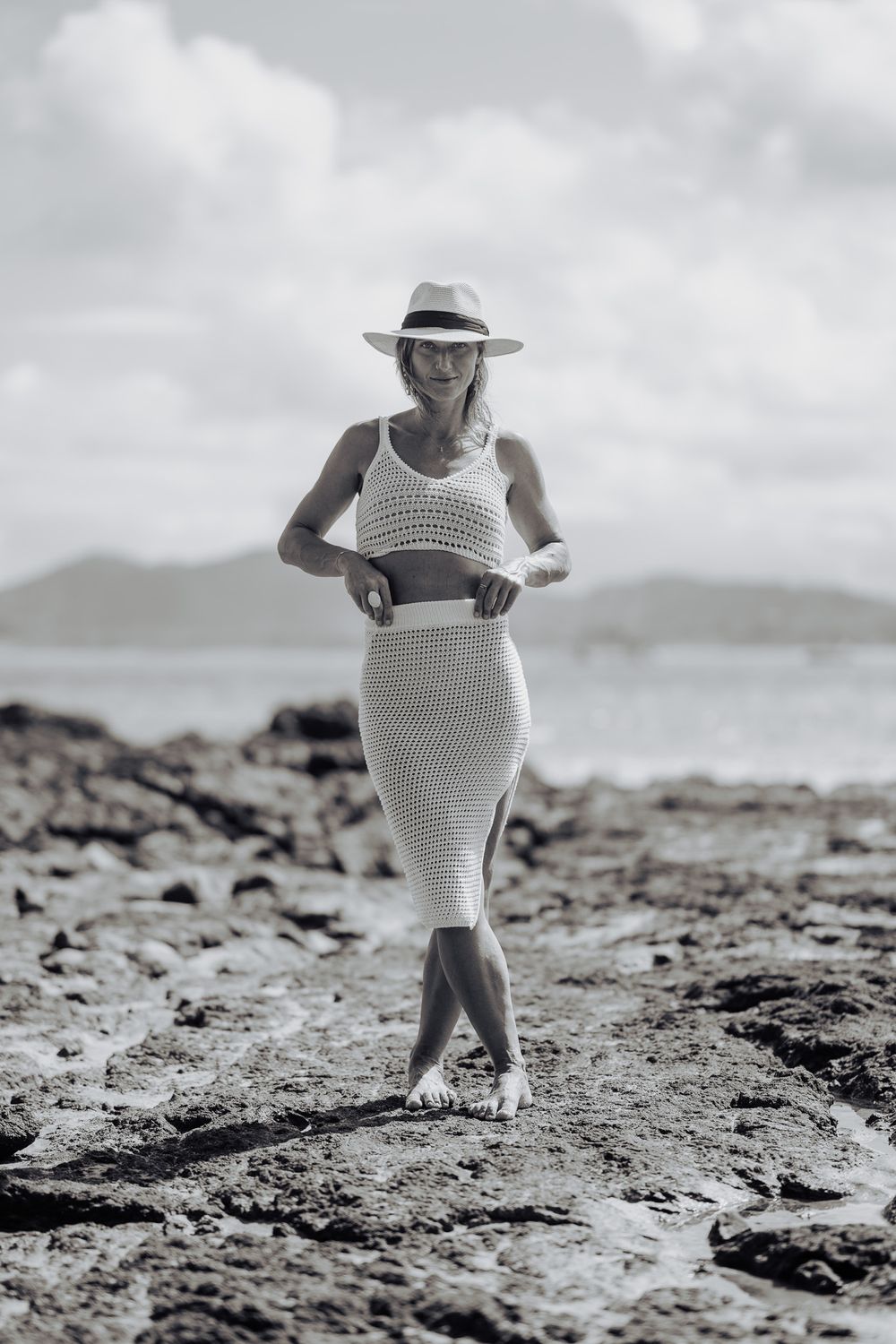 A black and white photo shows a figure in patterned dress exploring a rocky beach landscape with distant mountains.