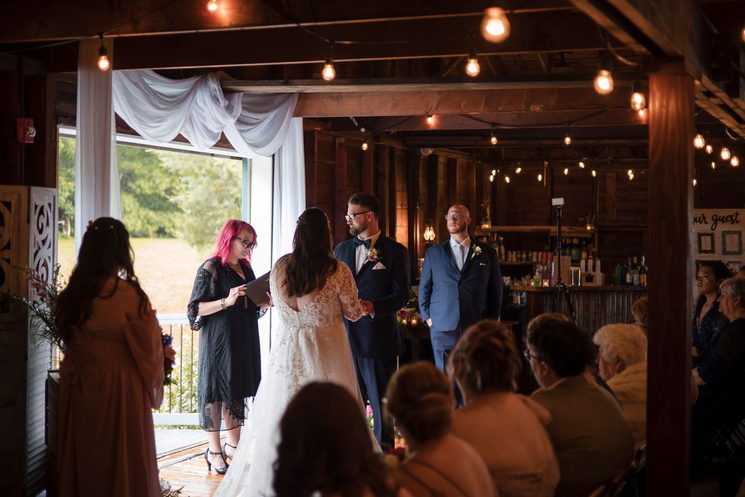 Wedding ceremony taking place in rustic barn venue with string lights and wooden beams overhead as guests look on.