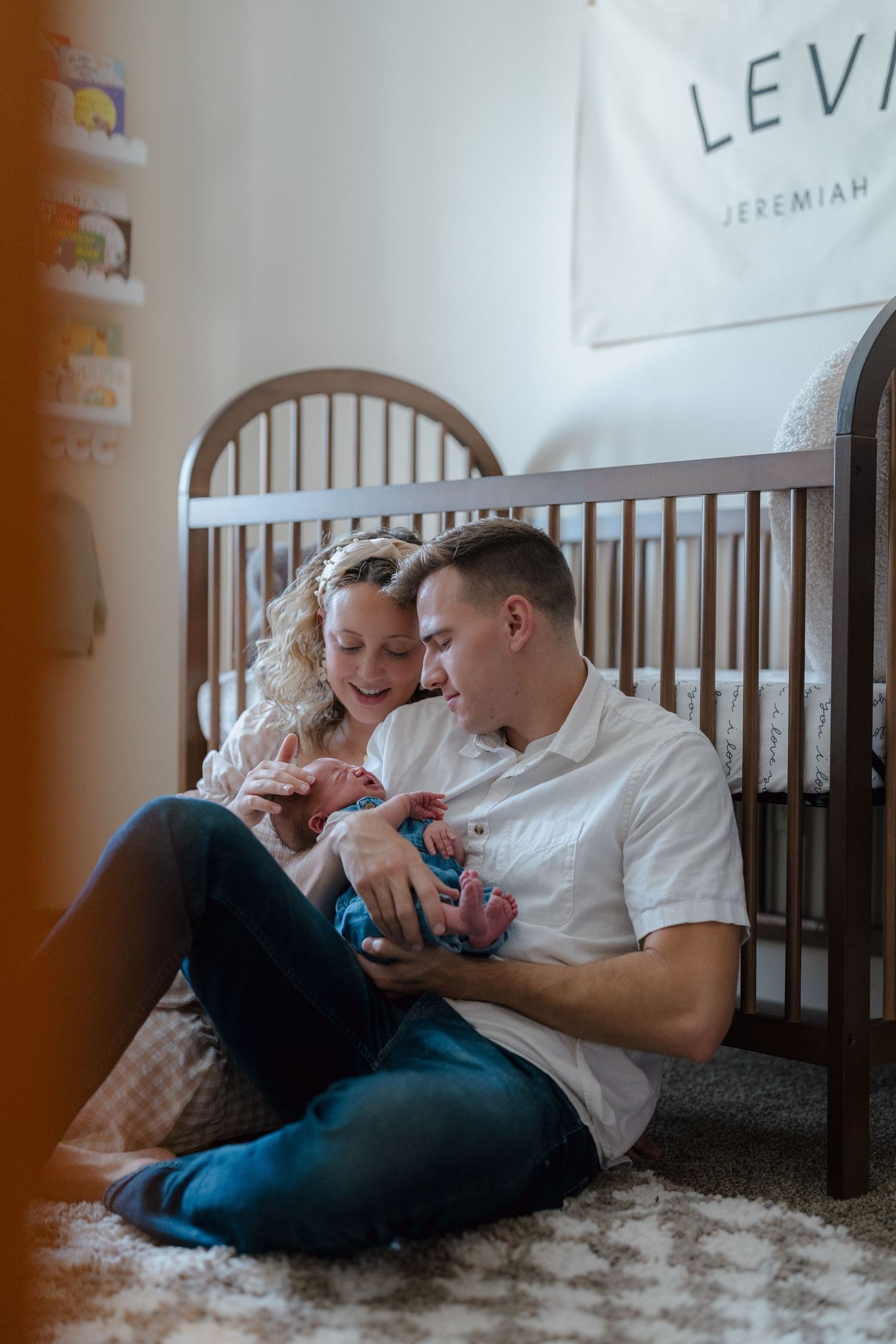 Two parents sit on a plush rug next to a wooden crib, tenderly holding their newborn baby in a cozy nursery setting.