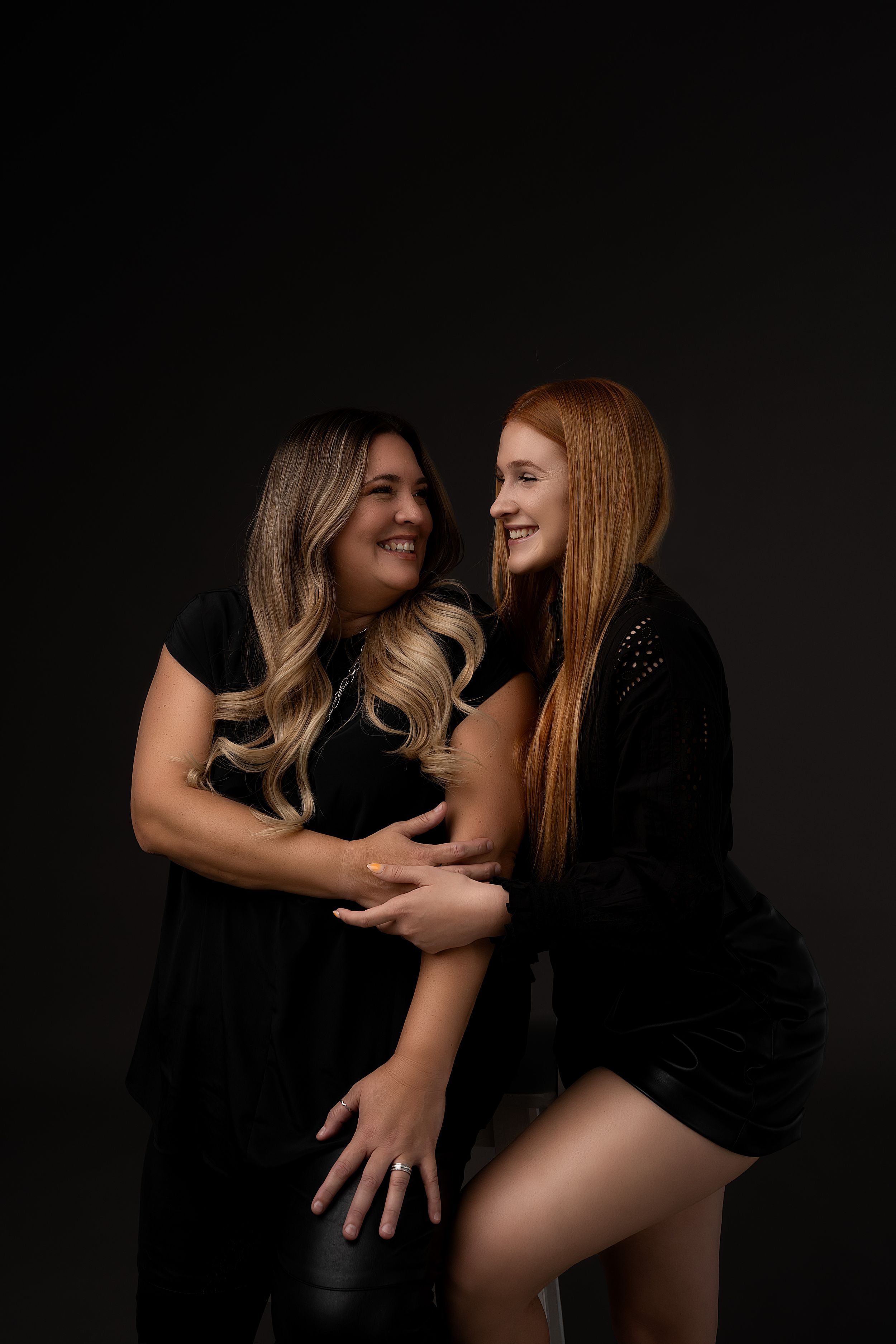 Two women in black outfits share a joyful moment together against a dark background during a studio photoshoot.