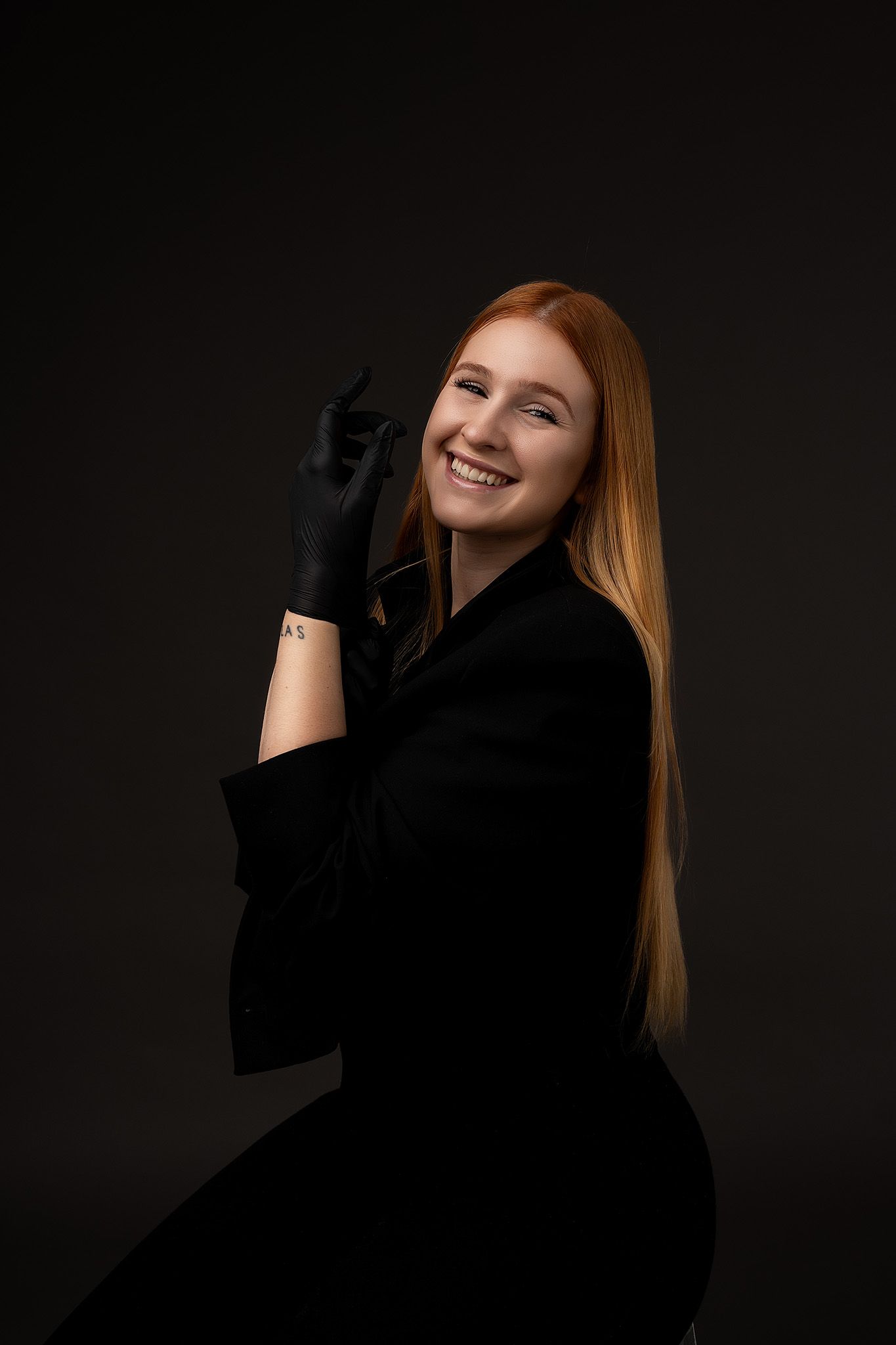 Portrait on dark background featuring someone in black clothing with long red hair posing while wearing dark gloves.
