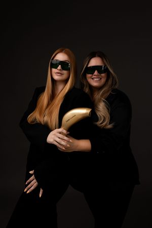 Two smiling people in black clothing and sunglasses pose for a series of fashionable portraits against a dark background.