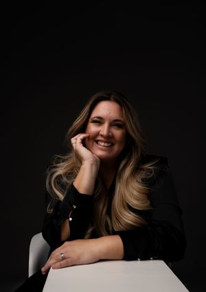 Professional studio portrait against dark background showing a warm smile and casual pose while wearing a black blazer.