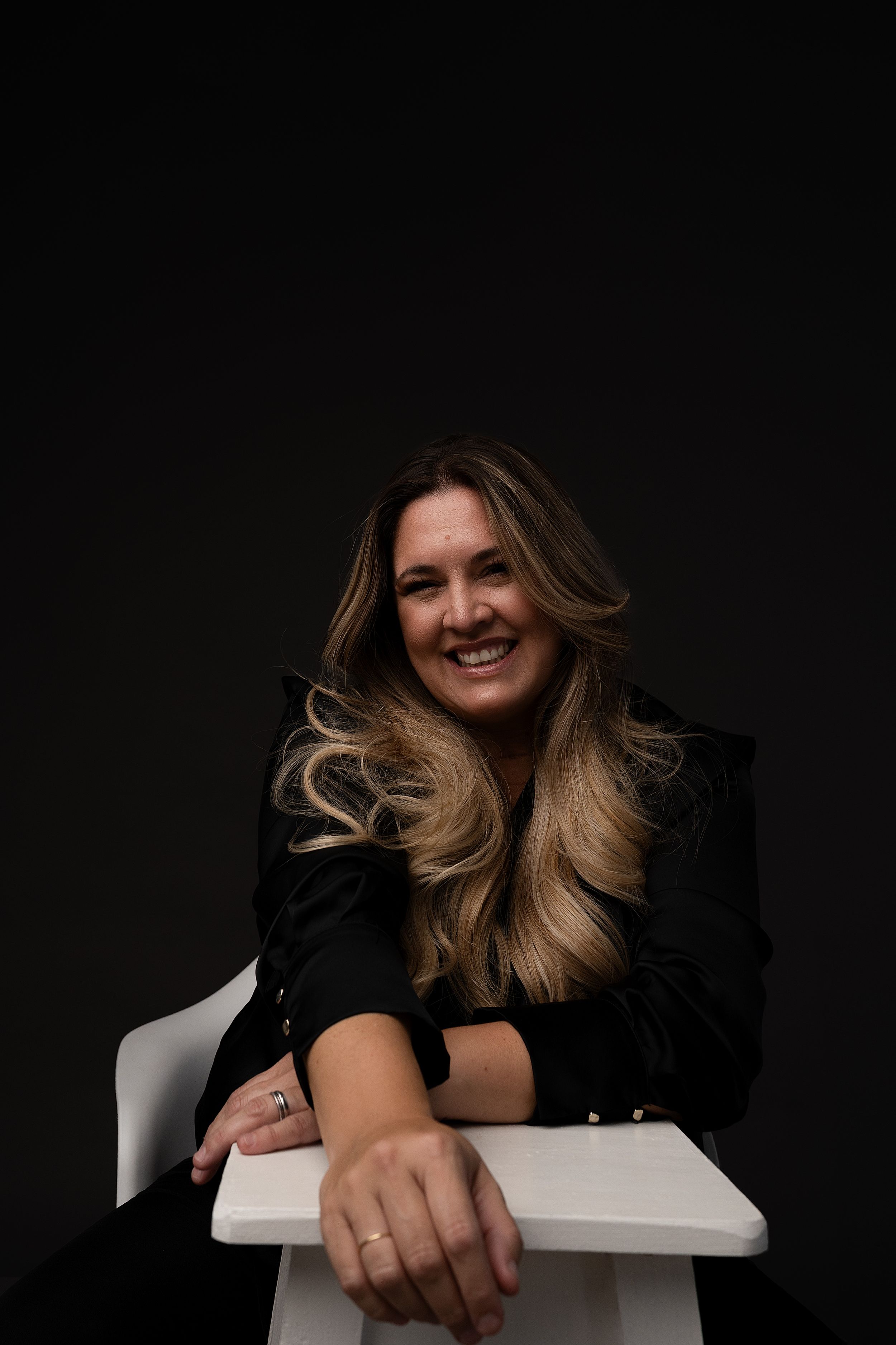 A woman in a black top leans over a white chair against a dark background with dynamic movement in her blonde hair.