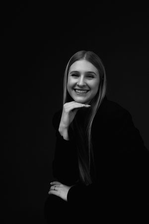 Black and white portrait of someone with long straight hair wearing a black sweater smiling against dark background.