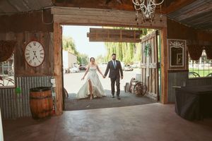 Couple walking through rustic barn doorway with willow trees visible in background.
