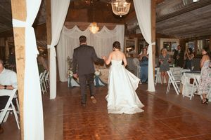 Couple walking hand in hand across wooden dance floor during reception with white draping and chandeliers.
