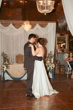 A couple shares their first dance at a rustic wedding reception under crystal chandeliers and elegant white draping.