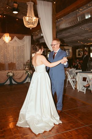 Couple shares a dance on wooden floor beneath crystal chandeliers and draped white fabric at wedding reception.