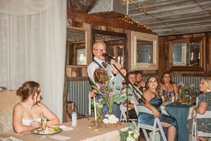 A speaker delivers a toast during a wedding reception in a rustic venue with string lights and wooden walls.