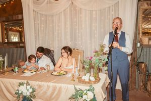 Wedding reception setting with head table and speaker addressing guests in rustic barn venue.
